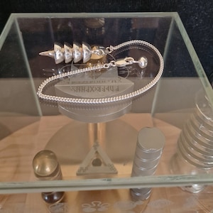 May include: A silver pendulum with a conical shape and a chain. The pendulum is in a glass display case with a wooden base. The chain is attached to a small metal ring. The pendulum is labeled "FIBERXPHRYS" and "V.A. CHRISTIANSEN".