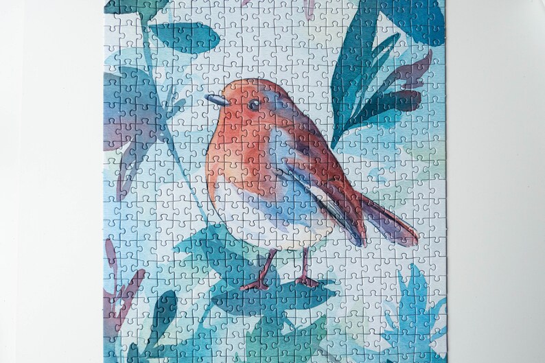 May include: A watercolor jigsaw puzzle featuring a robin perched on a branch surrounded by blue and green leaves.