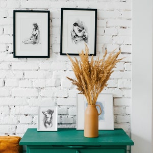 May include: A green dresser with six drawers against a white brick wall. Three black framed drawings of figures are hanging on the wall above the dresser. A vase with dried pampas grass is on top of the dresser.