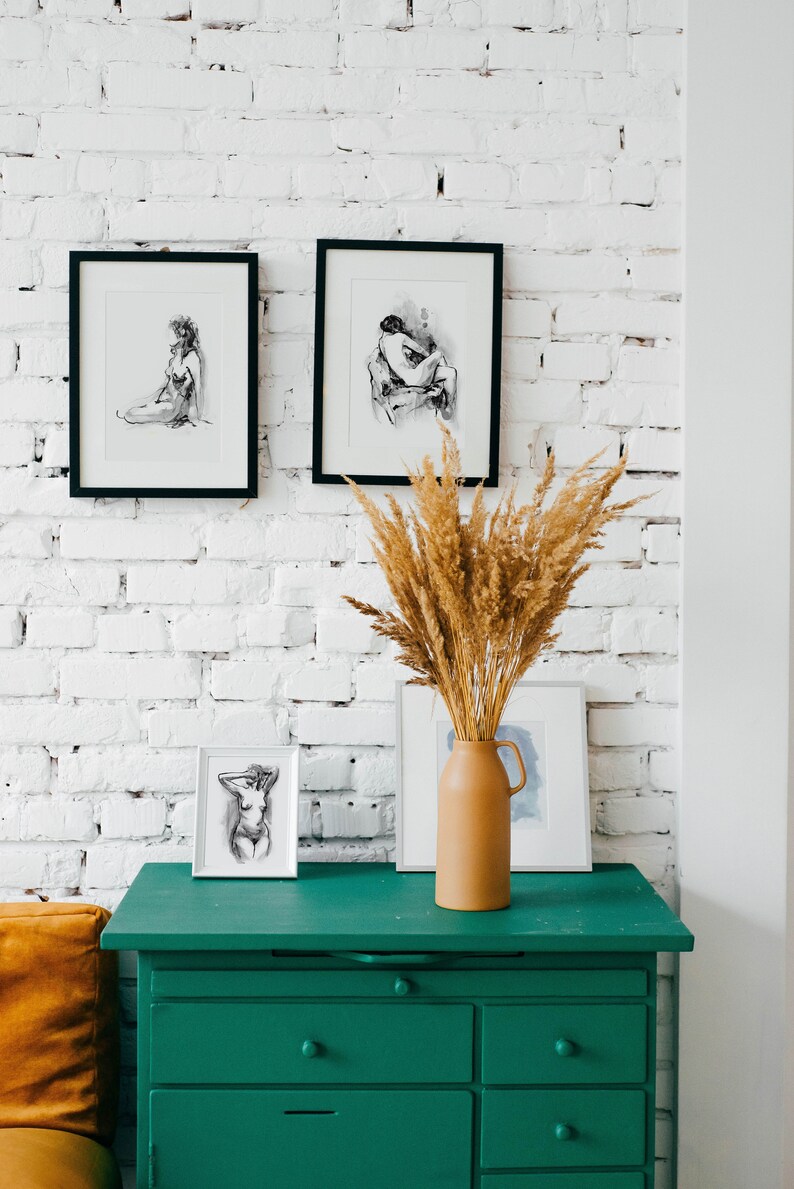 May include: A green dresser with six drawers against a white brick wall. Three black framed drawings of figures are hanging on the wall above the dresser. A vase with dried pampas grass is on top of the dresser.