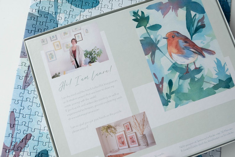 May include: A white box with a watercolor painting of a robin on a branch with green leaves. The box also features a photo of the artist and text that reads "Hel I am (ansal" and "I am so glad you got your arches pack, please enjoy!"