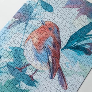 May include: A watercolor jigsaw puzzle featuring a robin perched on a branch with blue and green leaves. The bird is painted in shades of red, orange, and blue.