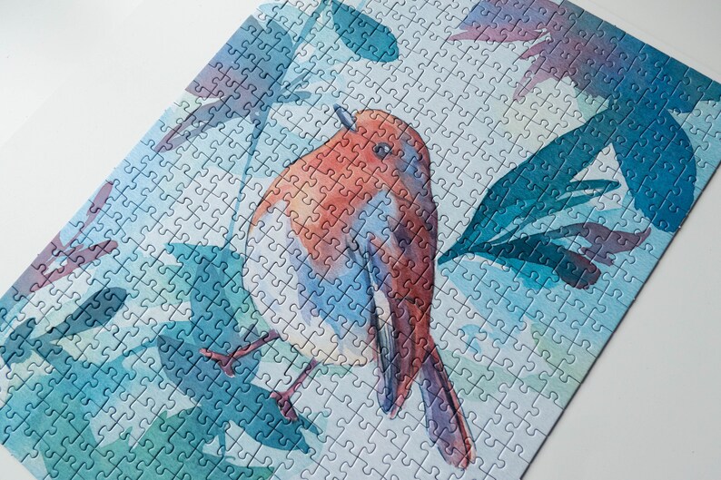 May include: A watercolor jigsaw puzzle featuring a robin perched on a branch with blue and green leaves. The bird is painted in shades of red, orange, and blue.