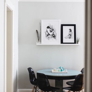 May include: A white wall with two black framed drawings of figures, a white shelf with a black feather, and a round glass table with four black chairs.