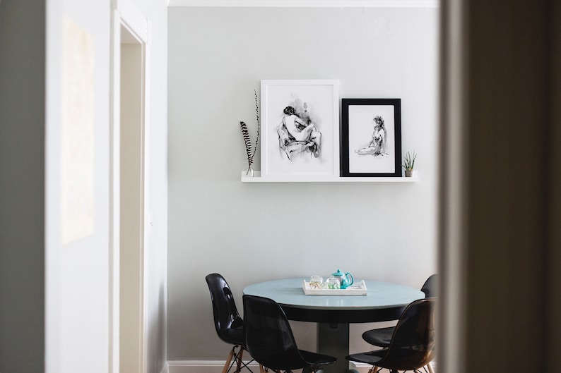 May include: A white wall with two black framed drawings of figures, a white shelf with a black feather, and a round glass table with four black chairs.