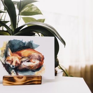 May include: A watercolor painting of a sleeping fox curled up on a blue and yellow background. The painting is on a white card and is displayed on a wooden easel.