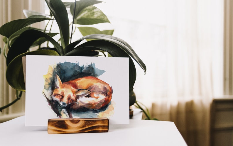 May include: A watercolor painting of a sleeping fox curled up on a blue and yellow background. The painting is on a white card and is displayed on a wooden easel.
