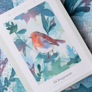 May include: A 50-piece jigsaw puzzle featuring a watercolor painting of a robin perched on a branch with green leaves. The puzzle box has the title "Ink Imaginarium" and the artist's name "Laura Jane McLaughlin".