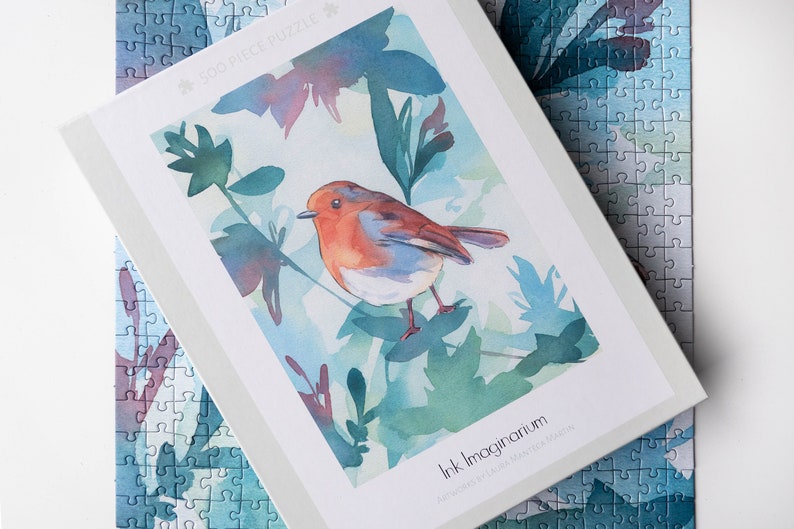 May include: A 50-piece jigsaw puzzle featuring a watercolor painting of a robin perched on a branch with green leaves. The puzzle box has the title "Ink Imaginarium" and the artist's name "Laura Jane McLaughlin".