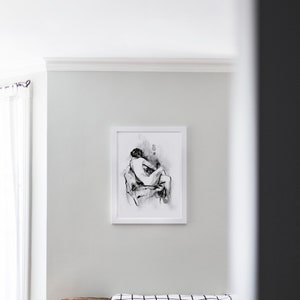 May include: A black and white framed print of a figure drawing of a person sitting with their arms wrapped around their legs. The print is hanging on a white wall above a brown couch with black and white patterned pillows.
