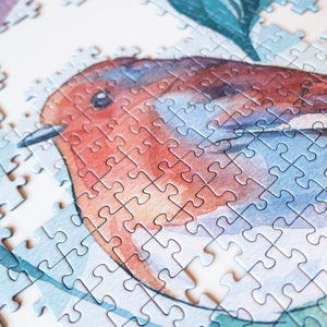 May include: A close-up of a completed jigsaw puzzle featuring a watercolor painting of a robin perched on a branch with green leaves. The puzzle pieces are small and intricate, creating a detailed and realistic image.