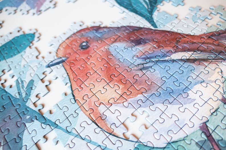 May include: A close-up of a completed jigsaw puzzle featuring a watercolor painting of a robin perched on a branch with green leaves. The puzzle pieces are small and intricate, creating a detailed and realistic image.
