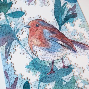 May include: A partially completed jigsaw puzzle featuring a watercolor painting of a robin perched on a branch with green leaves. The puzzle pieces are blue, green, and white.
