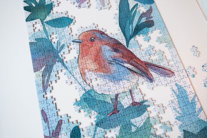 May include: A partially completed jigsaw puzzle featuring a watercolor painting of a robin perched on a branch with green leaves. The puzzle pieces are blue, green, and white.