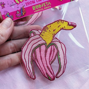 May include: Pink and yellow iron-on patch of a finger banana with the text "Fingerbanana" and "Iron on Patch" in a pink and yellow font. The text "ZÖB/ZETA" and "@ONIRICAL_ZUBIETA" are also included in the image.