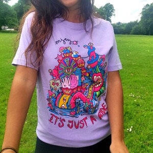以下が含まれることがあります： ラベンダー色のTシャツを着た女性。カラフルなグラフィックデザインが施されており、「enjoy... it's just a ride」と書かれています。ピンクのフープを持っています。