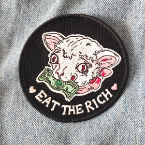 May include: Circular black patch featuring a white lamb's head with a green banknote in its mouth. The text "EAT THE RICH" is embroidered in white. Pink hearts flank the text. The patch is on denim.
