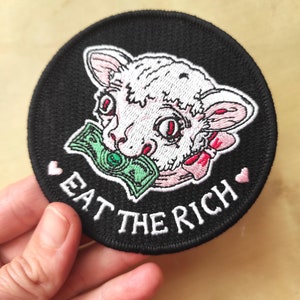 May include: Round black patch featuring a lamb with a dollar bill in its mouth. The lamb is white and pink with red eyes. The text "EAT THE RICH" is embroidered in white, with pink hearts on either side.