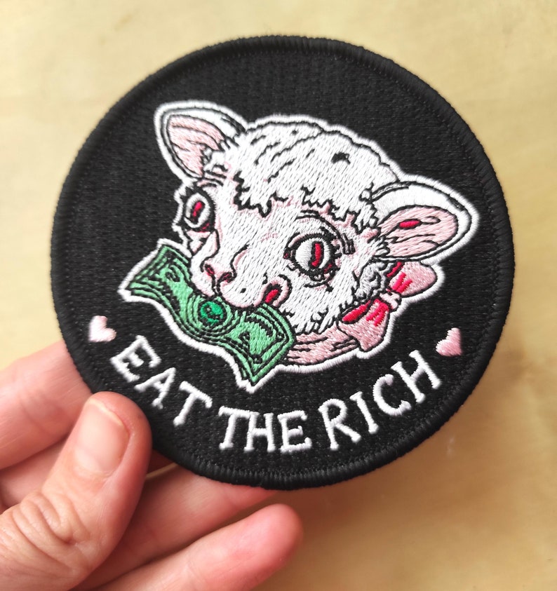 May include: Round black patch featuring a lamb with a dollar bill in its mouth. The lamb is white and pink with red eyes. The text "EAT THE RICH" is embroidered in white, with pink hearts on either side.
