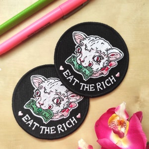 May include: Two black, round patches with a white cat design biting a dollar bill. The text "EAT THE RICH" is embroidered in white, with pink hearts. A pink and green marker are in the background.