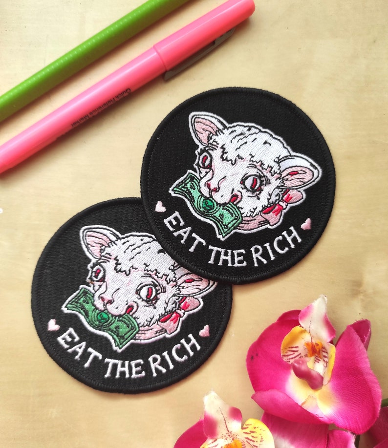 May include: Two black, round patches with a white cat design biting a dollar bill. The text "EAT THE RICH" is embroidered in white, with pink hearts. A pink and green marker are in the background.