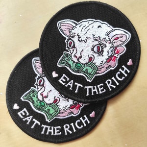 May include: Two round black patches with white embroidered text and a cartoon lamb design. The lamb holds a green bill in its mouth and is surrounded by pink accents. The text on the patches reads "EAT THE RICH."