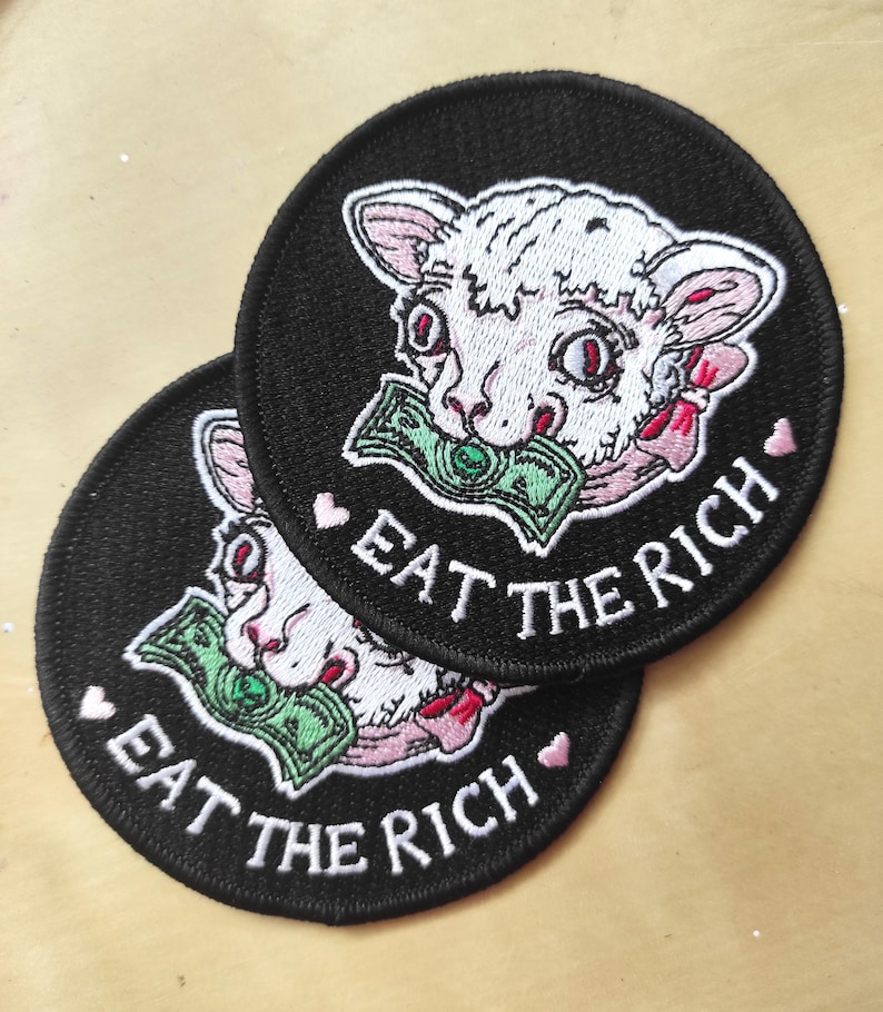 May include: Two round black patches with white embroidered text and a cartoon lamb design. The lamb holds a green bill in its mouth and is surrounded by pink accents. The text on the patches reads "EAT THE RICH."