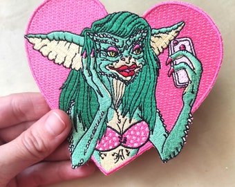 May include: A pink heart-shaped embroidered patch featuring a green-skinned creature with long green hair, wearing a pink bikini top and holding a phone. The creature has large ears and is looking at the phone.
