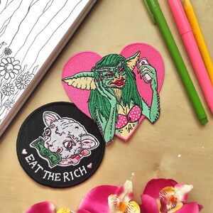 May include: An assortment of embroidered patches featuring pop culture and satirical designs. One patch depicts a Gremlin taking a selfie on a pink heart background. Another shows a cat with money in its mouth, with the text "EAT THE RICH".