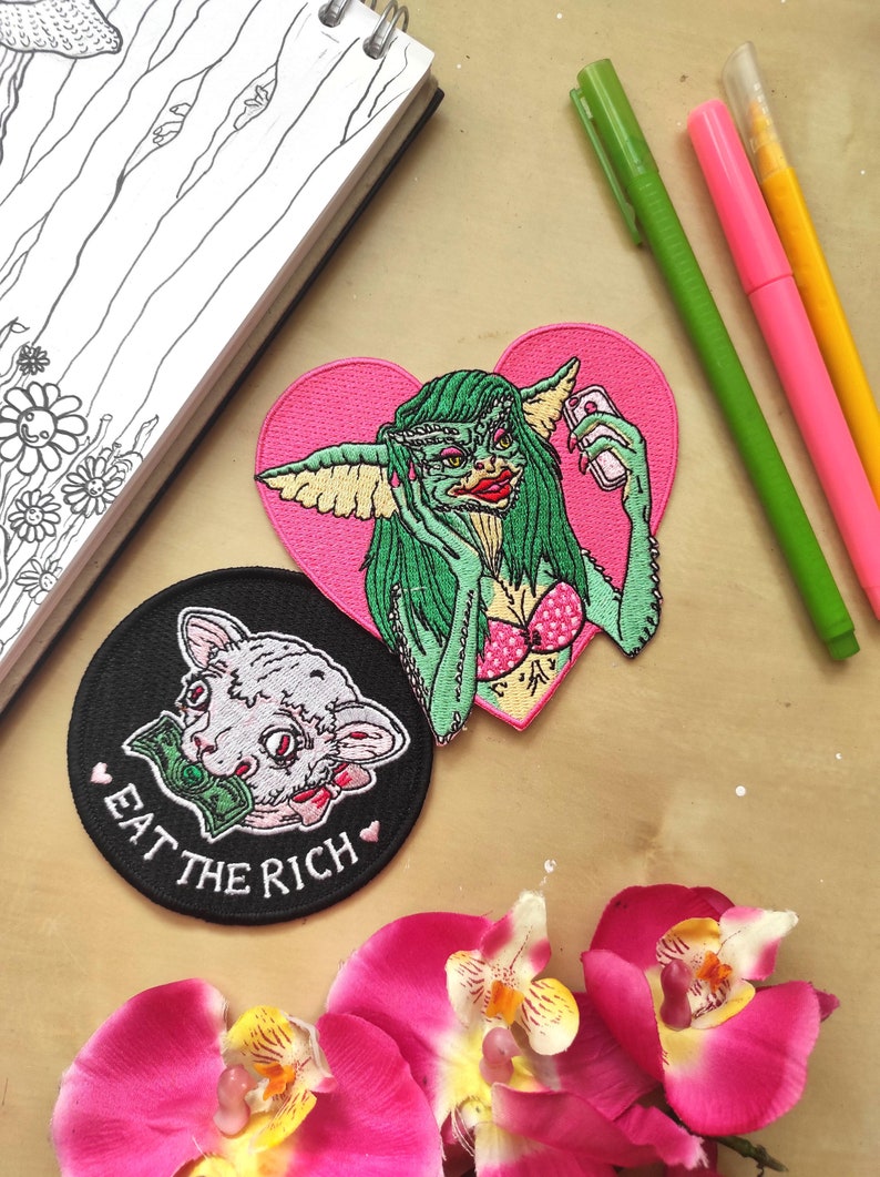 May include: An assortment of embroidered patches featuring pop culture and satirical designs. One patch depicts a Gremlin taking a selfie on a pink heart background. Another shows a cat with money in its mouth, with the text "EAT THE RICH".