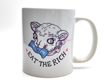 以下が含まれることがあります： 白いセラミック製のマグカップに、ドル札を噛んでいる羊の漫画のイラストが描かれています。羊の下には「Eat the Rich」という文字が印刷されています。