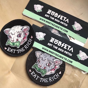 May include: Black, round embroidered patches with a cat graphic biting a wad of money, and the text "EAT THE RICH". The patches are next to black and white business cards with the text "ZUBIETA EAT THE RICH PATCH".