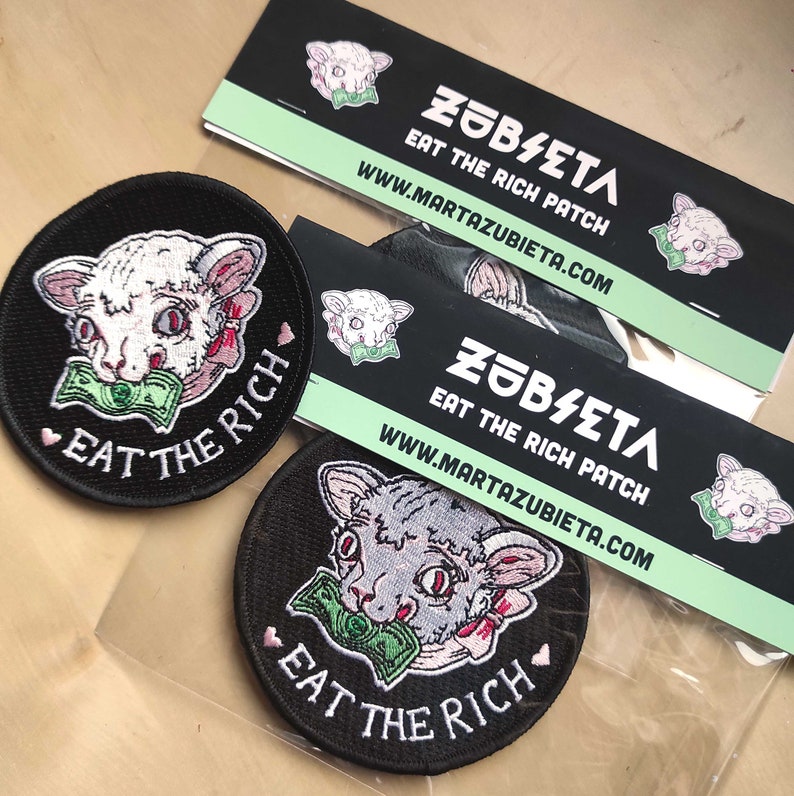 May include: Black, round embroidered patches with a cat graphic biting a wad of money, and the text "EAT THE RICH". The patches are next to black and white business cards with the text "ZUBIETA EAT THE RICH PATCH".