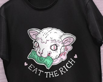 以下が含まれることがあります： 黒いTシャツに、白い漫画の羊が緑色の紙幣を食べており、ピンク色の文字で「Eat the Rich」と書かれている。