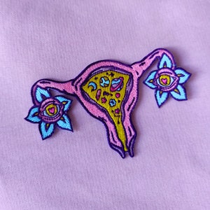 May include: A pink and yellow embroidered patch of a uterus with flowers and celestial bodies inside. The flowers have blue petals and a pink center with a heart and eye.