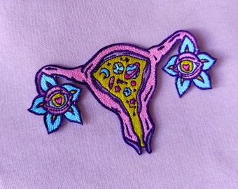 May include: A pink and yellow embroidered patch of a uterus with flowers and celestial bodies inside. The flowers have blue petals and a pink center with a heart and eye.