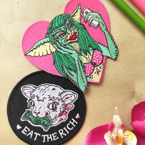 May include: Embroidered patches featuring a green creature with a pink heart background, holding a phone, and a white cat with money in its mouth on a black circle patch with the text "EAT THE RICH".