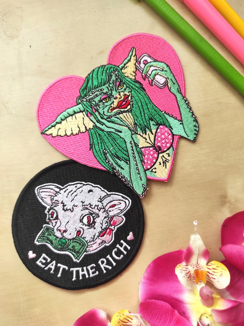 May include: Embroidered patches featuring a green creature with a pink heart background, holding a phone, and a white cat with money in its mouth on a black circle patch with the text "EAT THE RICH".