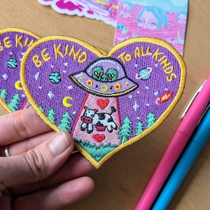 May include: Heart-shaped embroidered patch with the text "BE KIND TO ALL KINDS". The patch features a purple background with a UFO, aliens, a cow, trees, planets, and stars. The patch has a yellow border.