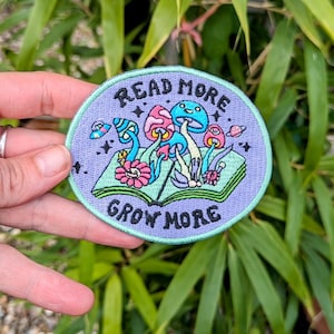 May include: A circular embroidered patch with a pale purple backdrop. It depicts vibrant mushrooms sprouting from an open book, with the words "READ MORE" and "GROW MORE" in black text. The patch is held by a hand.