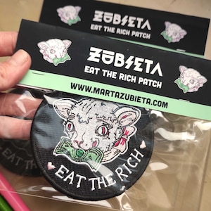 May include: A black round patch with the text "EAT THE RICH" and a cartoon cat with money in its mouth. The patch is packaged with a black card that has the text "ZUBIETA EAT THE RICH PATCH" and the website address.