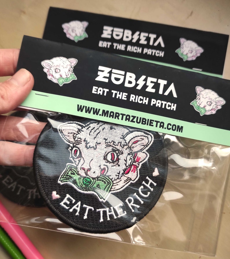 May include: A black round patch with the text "EAT THE RICH" and a cartoon cat with money in its mouth. The patch is packaged with a black card that has the text "ZUBIETA EAT THE RICH PATCH" and the website address.