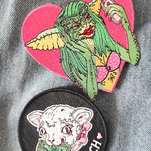 May include: Two embroidered patches on denim. One is heart-shaped with a green creature holding a phone. The other is round, black, with a lamb holding money and the text "EAT THE RICH".