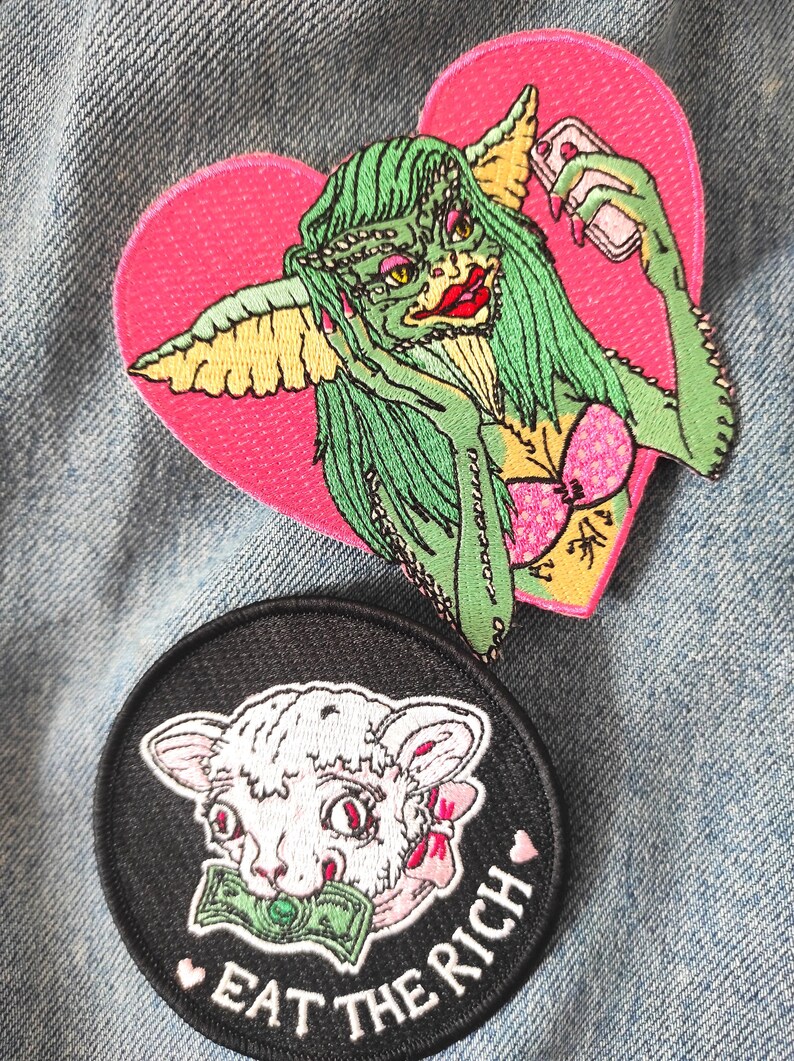 May include: Two embroidered patches on denim. One is heart-shaped with a green creature holding a phone. The other is round, black, with a lamb holding money and the text "EAT THE RICH".