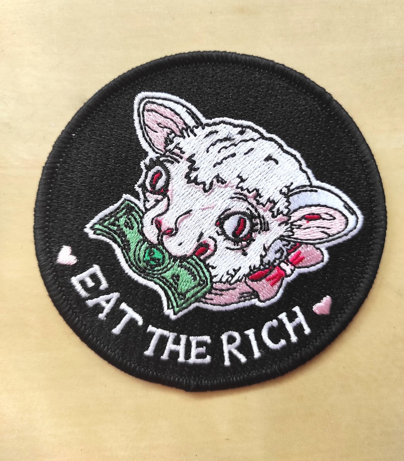 May include: Embroidered patch with a black background featuring a white cat-like creature with red eyes and a green dollar bill in its mouth. The text "EAT THE RICH" is in white, with pink hearts.