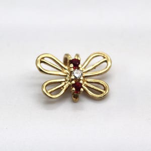 May include: A gold butterfly pendant featuring open-work wings and a central body set with two round red gemstones flanking a single clear round gemstone in the middle. The piece has a small bail at the top for attachment to a chain or cord.