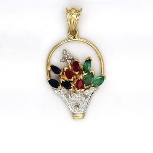 May include: A gold pendant shaped like a basket filled with marquise-cut gemstones. The arrangement includes blue sapphires, red rubies, and green emeralds set within a circular gold frame. The basket base features a textured silver-tone finish.