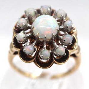May include: A gold ring featuring a large, round opal at the center, surrounded by smaller opals. The opals have a milky white appearance with hints of color. The ring's setting has a detailed, ornate design.