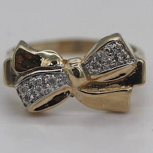 May include: A gold-toned ring in a bow shape, with a cluster of small diamonds set in the center of each bow loop. The ring has a polished finish and a classic design, suitable for everyday wear or special occasions.