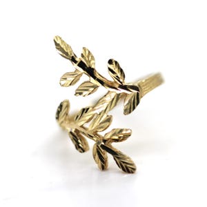 May include: A gold ring featuring an open design with two overlapping leaf-shaped branches. The metal surface of the leaves has a textured, faceted finish that reflects light, creating a shimmering appearance across the entire piece of jewelry.